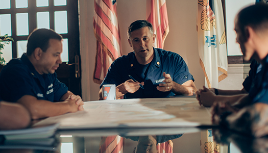 Military personnel are seated at a large table with a map on it having a conversation.