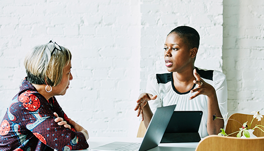 Two women are seated at a table with one of them explaining something to the other.